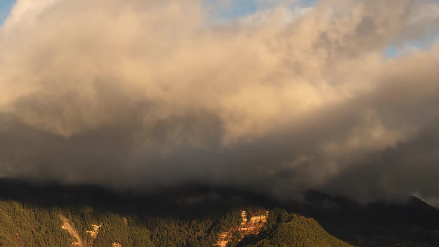An aerial view of golden hour clouds rolling over a lush, green mountain range, casting dramatic shadows across the landscape