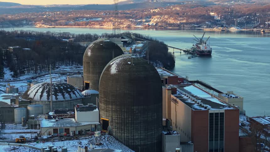 An aerial view orbits the Indian Point Nuclear Power Plant on the Hudson River during a snowy winter sunrise as a cargo ship floats nearby