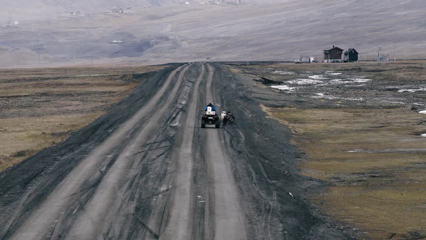 A person on an ATV with a team of sled dogs travels down a long, desolate road in a vast, barren landscape