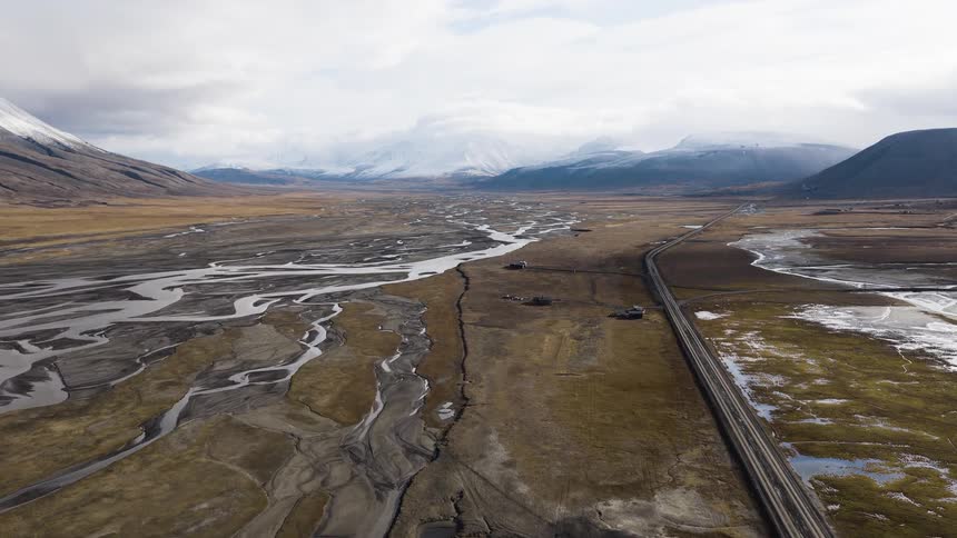 A sweeping aerial view reveals a vast, barren valley with a braided river system and a long, straight road leading towards distant snow-capped mountains