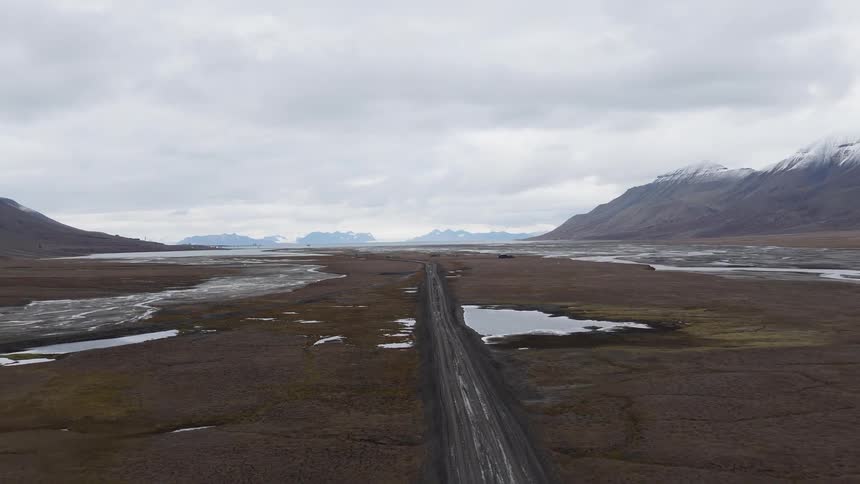 An aerial view follows a long, remote dirt road through a vast, barren valley flanked by snow-capped mountains in a desolate arctic landscape
