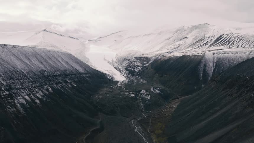 A cinematic aerial view slowly reveals a majestic, snow-dusted mountain valley and a distant glacier under a cloudy sky