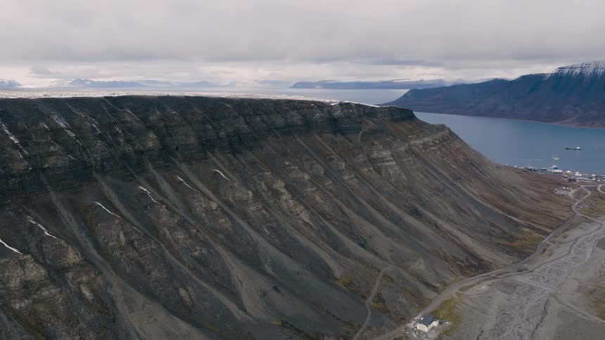 A slow, dramatic aerial push-in towards a vast, layered mountain overlooking a remote arctic town nestled in a fjord
