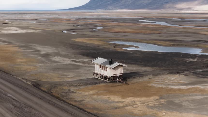 A slow aerial orbit reveals a solitary wooden cabin in a vast, desolate Arctic landscape near a quiet road and fjord
