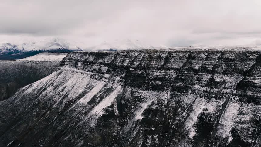A drone slowly ascends over a majestic, snow-covered cliff revealing a vast, frozen mountain landscape