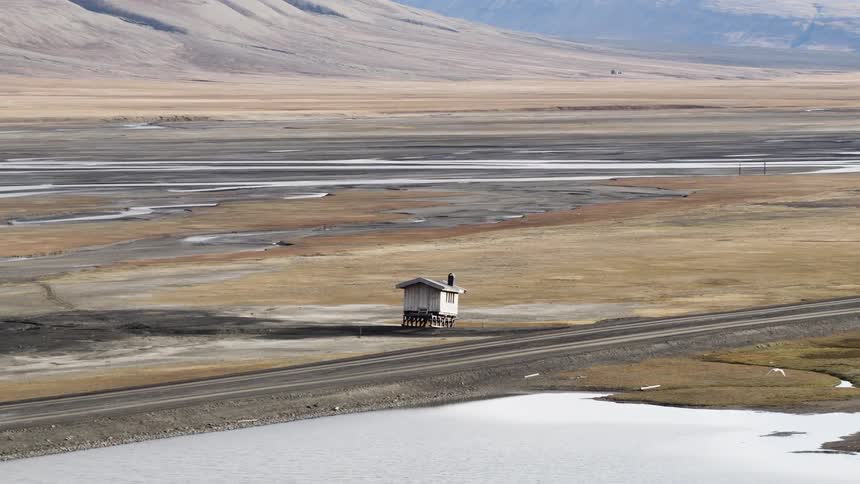 A slow zoom focuses on a solitary cabin on stilts amidst a vast, desolate arctic tundra landscape