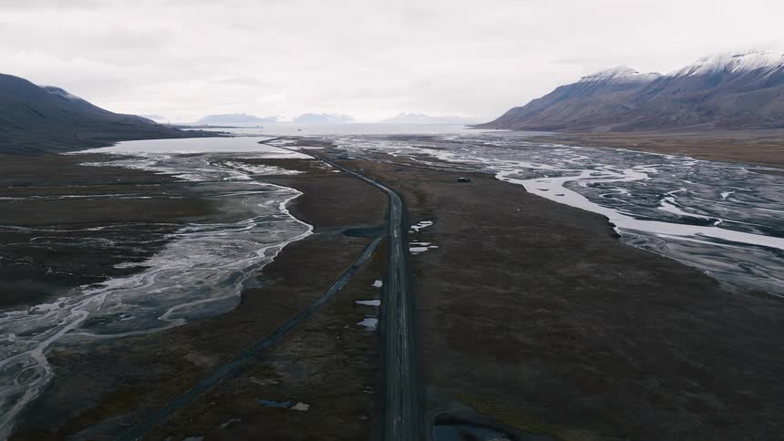 An aerial drone shot slowly flies over a desolate arctic valley with a long road alongside a frozen river leading to the ocean