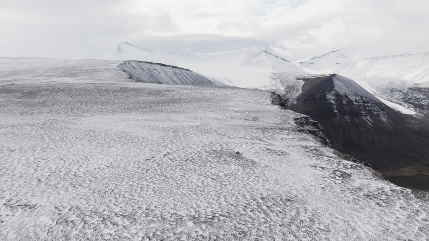 An expansive aerial shot slowly pulls back over a vast, snow-covered landscape revealing a desolate mountain range under a cloudy sky
