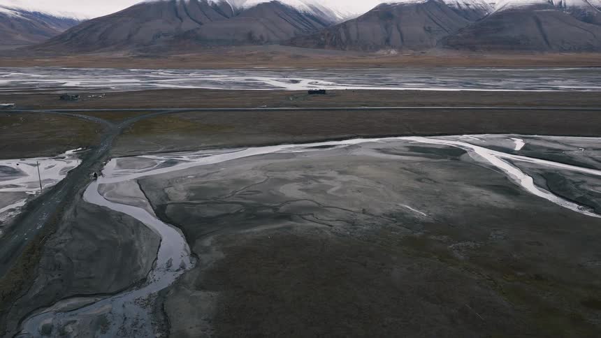 An aerial flight moves over a desolate arctic valley with a winding river and road, revealing a vast snow-capped mountain range
