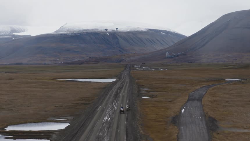 Two all-terrain vehicles travel down a long, desolate dirt road in a vast arctic valley surrounded by snow-dusted mountains