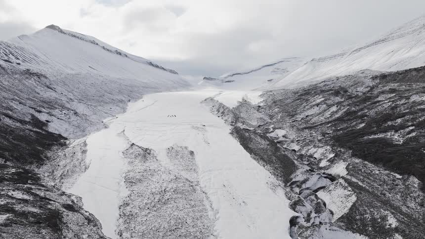 Stunning aerial view of a huge, snow-covered glacier flowing through a desolate mountain valley as tiny hikers traverse its surface