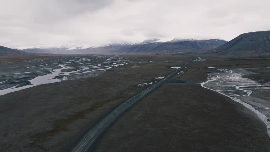 A slow, rising aerial shot reveals a vast, desolate arctic valley with a long, empty road leading to distant snow-capped mountains