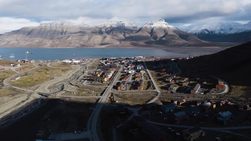 An aerial view pushes towards the remote arctic town of Longyearbyen in Svalbard, nestled between snow-capped mountains and a tranquil fjord