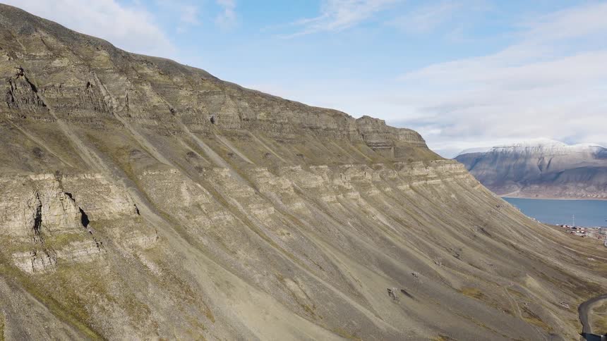 Slow aerial pullback shot of a vast, rocky mountainside towering over a remote coastal town in a majestic Arctic landscape
