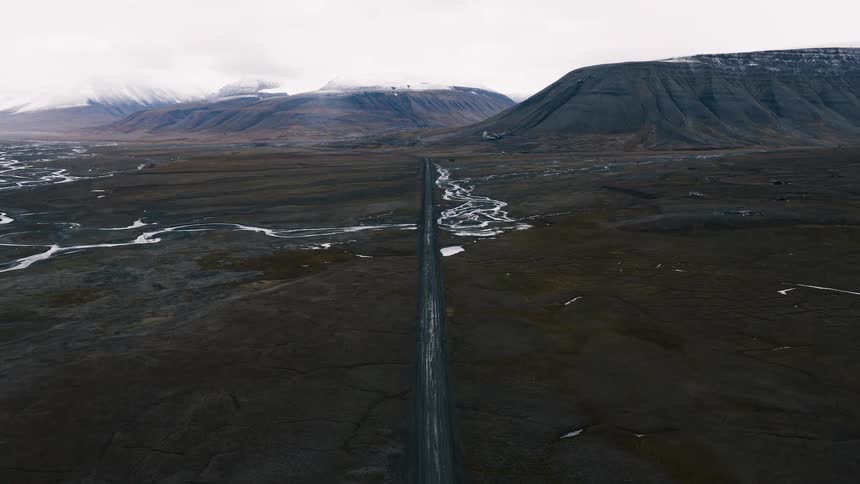 A high aerial shot follows a long, straight road through a desolate, vast valley with snow-capped mountains in the distance