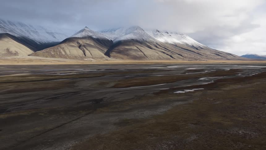 A slow aerial flyover reveals a lonely cabin in a vast arctic tundra, dwarfed by imposing snow-covered mountains
