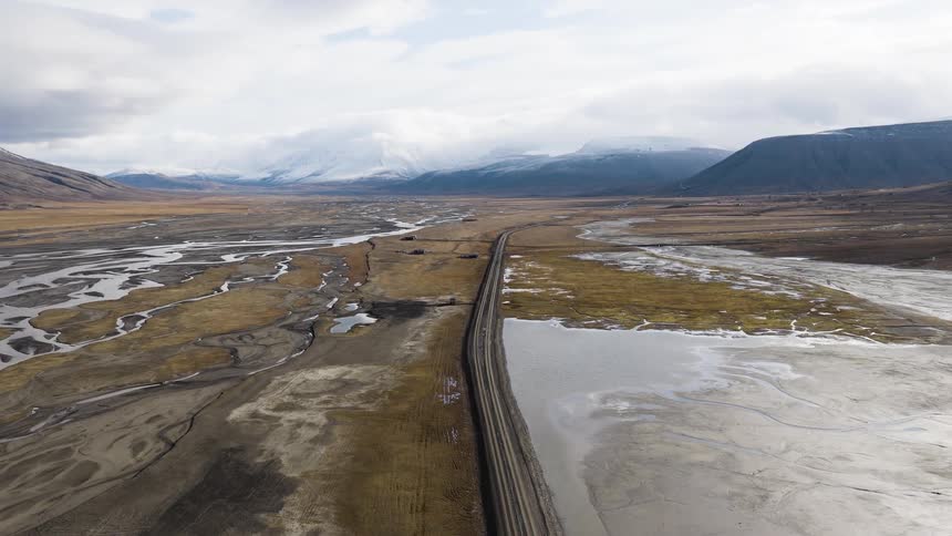 An aerial shot slowly pulls back over a long, straight road that divides a braided river and a partially frozen lake in a vast arctic valley with snow-capped mountains