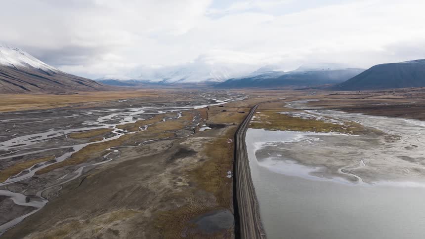 Epic aerial shot over a remote road dividing a braided river delta and a lake in a vast valley with snow-capped mountains in the distance