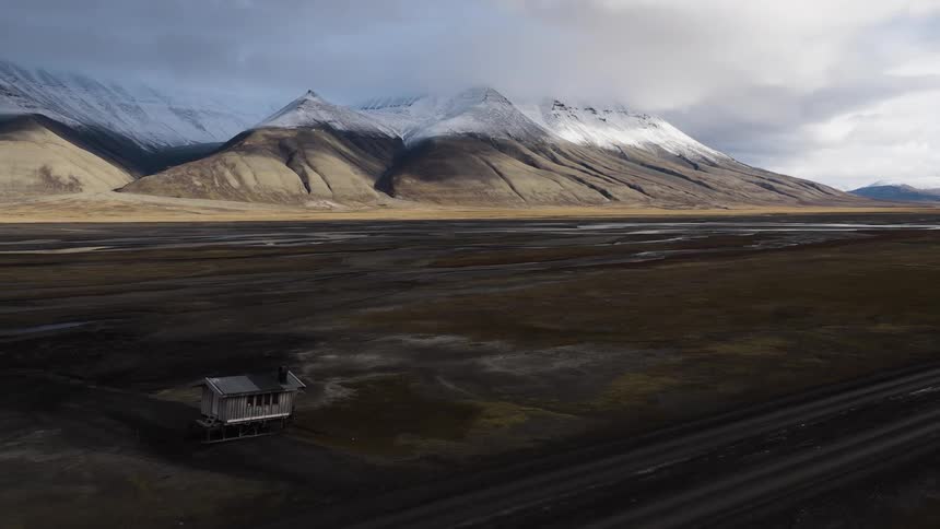 An epic aerial pullback over a vast, desolate river valley reveals a lone cabin against a backdrop of majestic, snow-capped mountains