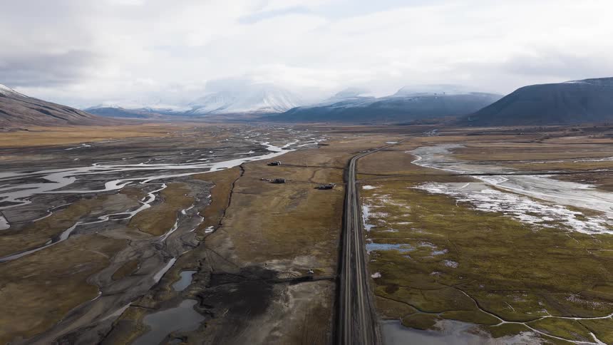 A slow aerial pullback reveals a vast, remote valley with a straight road alongside a braided river, leading towards distant snow-capped mountains