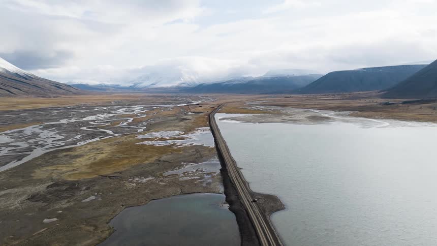 An aerial view of a remote road splitting a vast, open valley between a serene lake and a river delta, with snow-capped mountains in the distance