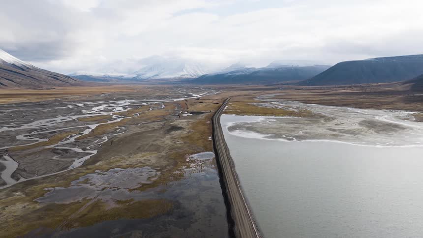 An aerial pullback reveals a long, straight road separating a vast river delta from a calm lake in a remote mountain valley