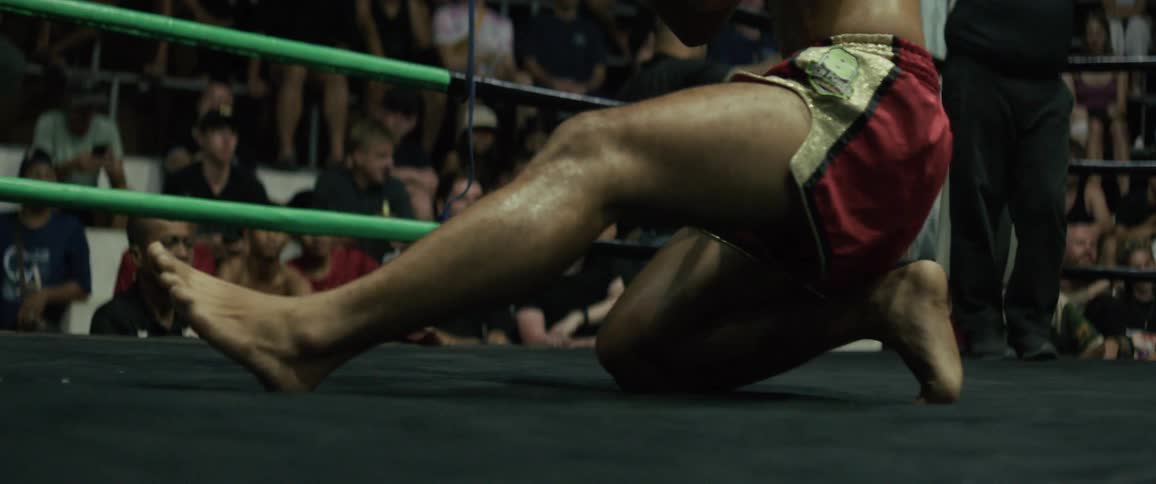 A low-angle slow-motion shot captures the muscular legs of a Muay Thai fighter performing a traditional pre-fight ritual in the ring