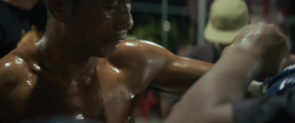 A sweaty, muscular Asian man grimaces as he receives an intense shoulder and back massage outdoors at night