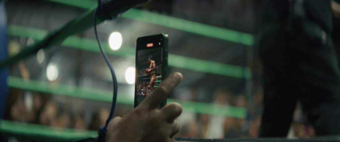 A spectator's hand holds a smartphone, capturing a live boxing match from the edge of the ring