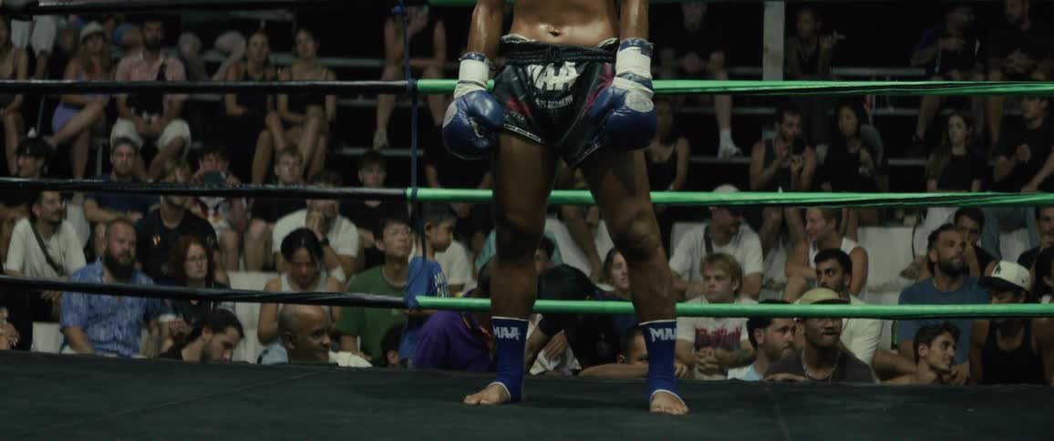 A sweaty and muscular Muay Thai fighter stands in the center of a boxing ring at night, looking intense and focused