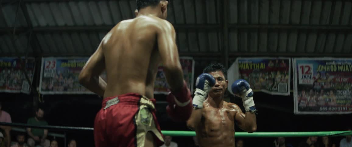 An intense, slow-motion close-up of two Muay Thai fighters grappling and trading blows in a dimly lit boxing ring