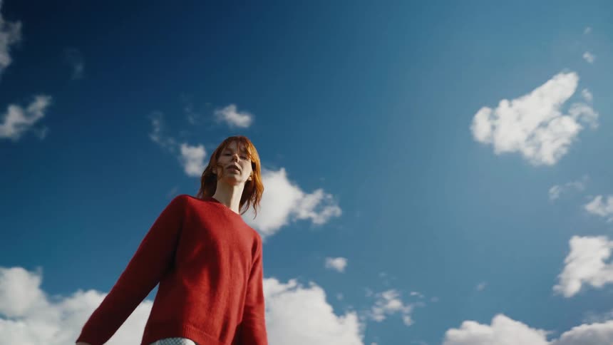 A stylish woman with flowing red hair walks and dances gracefully in slow motion against a dramatic blue sky with clouds