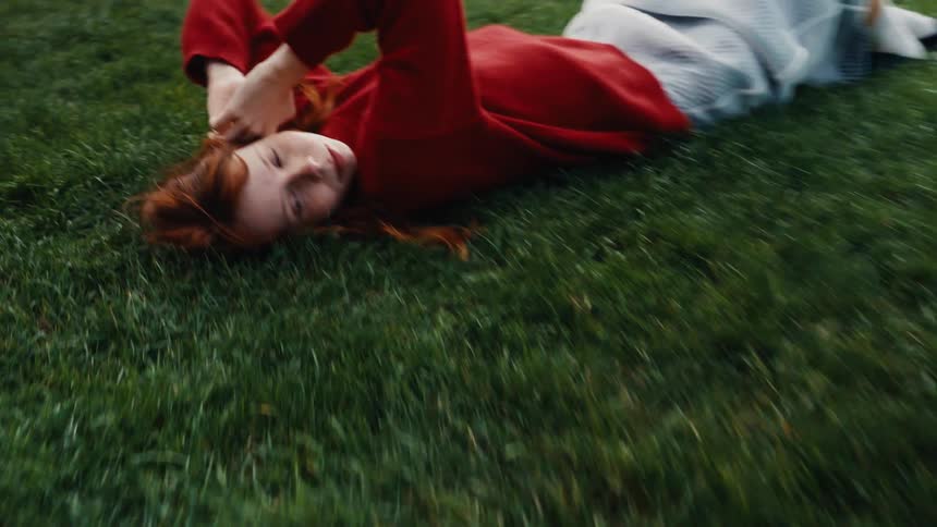 A camera glides over a lush green lawn, settling on a peaceful young redhead in a red sweater lying on her back