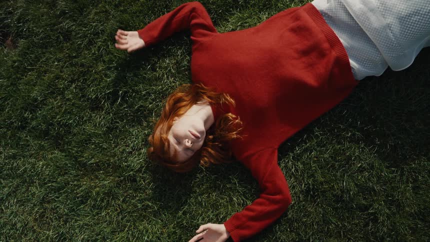 A top-down view shows a young woman with red hair slowly lying back to rest on the green grass in the sunshine