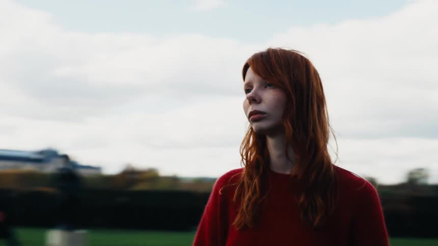 A beautiful woman with red hair walks through a park in Paris as the camera cinematically orbits around her, creating a dreamy and pensive mood