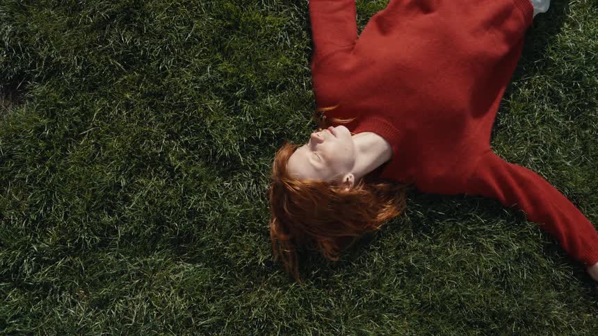 A serene red-haired woman in a matching sweater rests peacefully on the green grass in a spinning overhead shot
