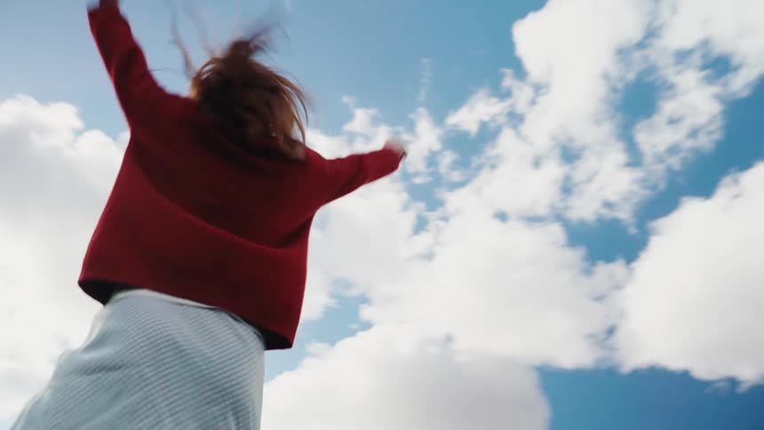 A woman with red hair dances expressively in slow motion under a bright blue sky filled with clouds, as seen from a low angle