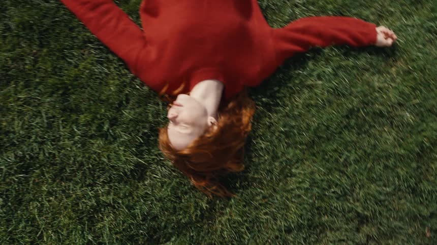 Overhead shot pans over a young woman with red hair relaxing peacefully in the green grass on a sunny day