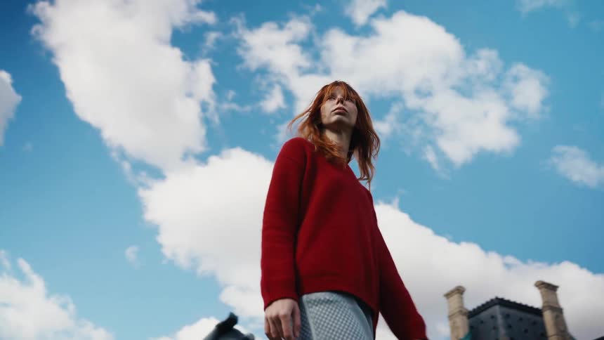 A pensive red-haired woman in a red sweater stands against a dramatic blue sky as the wind blows her hair in slow motion