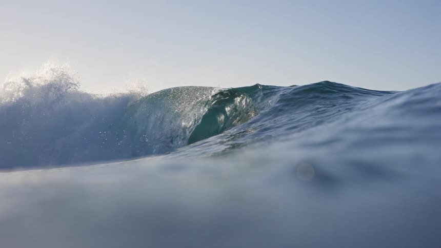 A stunning slow-motion, water-level shot shows a powerful ocean wave curling and breaking under a bright, clear sky