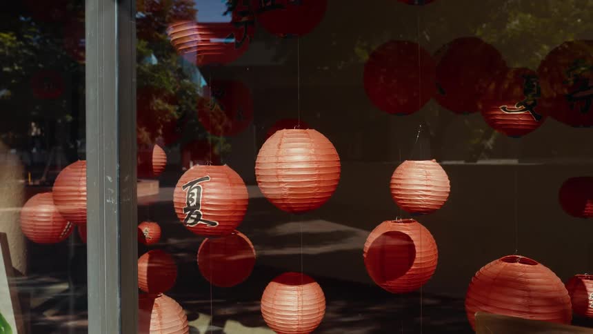 Red Japanese paper lanterns hang in a storefront window display, reflecting a quiet city street on a sunny day