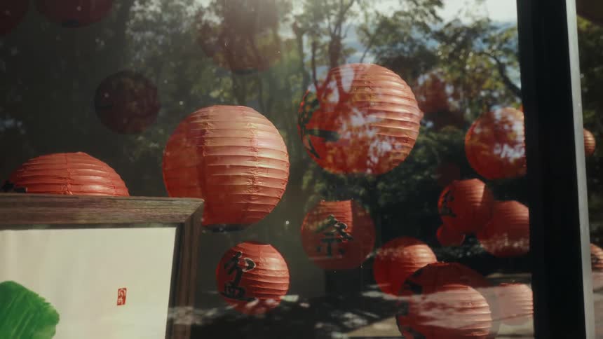 Sunlight streams through a window decorated with red paper lanterns as trees are reflected in the glass