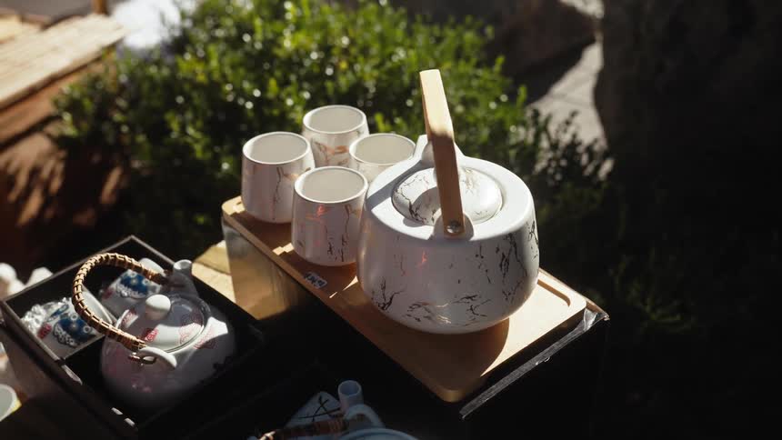 A modern white and gold marble-patterned ceramic tea set is displayed for sale outdoors in a sunny garden
