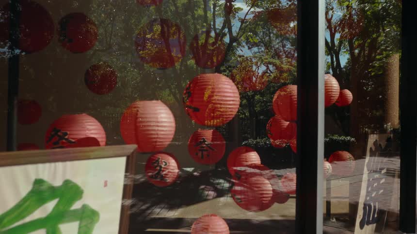 Red paper lanterns with Japanese characters hang in a window reflecting lush trees on a sunny day