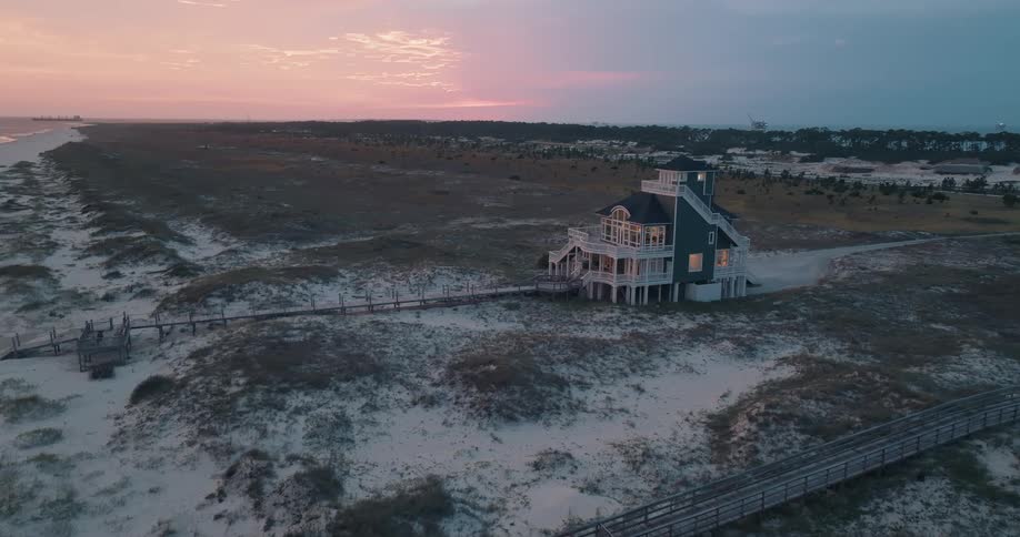 A still aerial shot captures a modern beach house on the dunes at sunset with lights glowing from within