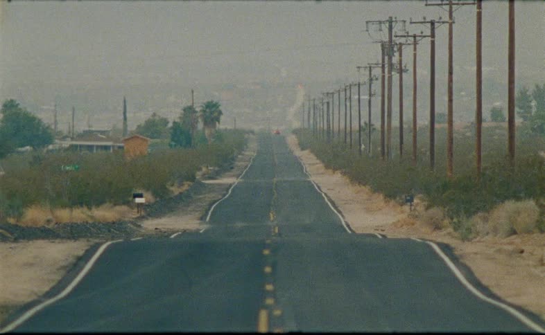A long, empty desert highway stretches into the hazy distance with telephone poles lining the side