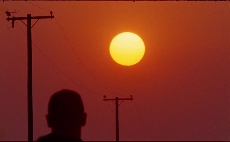 The silhouette of a person walks in front of telephone poles against the backdrop of a large, bright sun during sunset