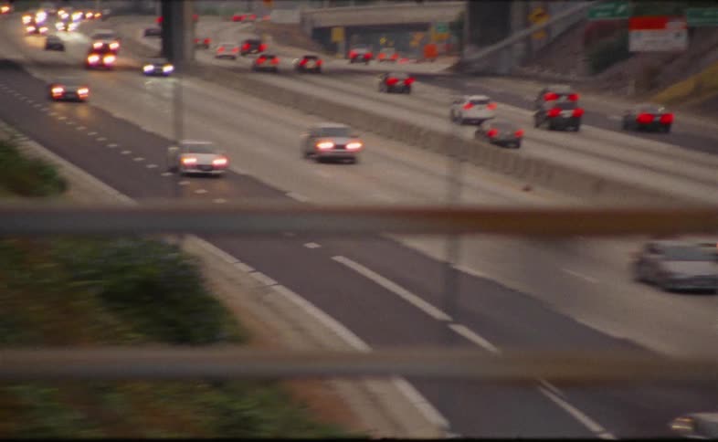 A focus pull reveals cars with headlights on driving along a busy Los Angeles freeway at dusk, seen through a fence