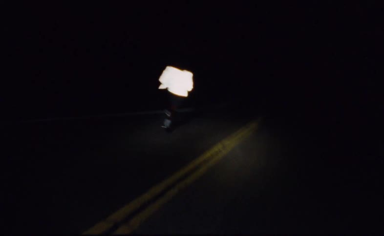 From behind, a man in a reflective jacket runs down a dark, empty road at night, illuminated by a single light