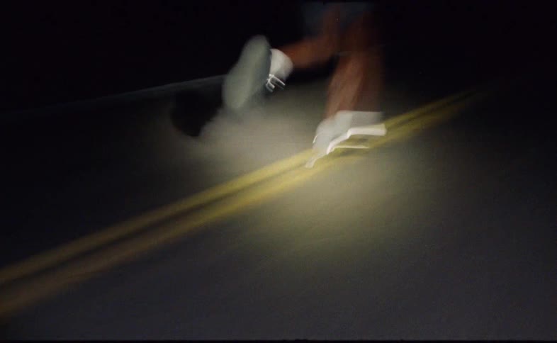 Close-up motion blur captures a runner's feet pounding the pavement of a dark road at night, illuminated by a single light